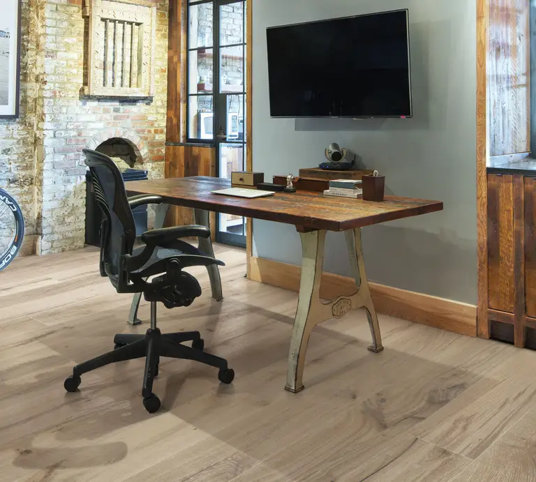 A cozy, industrial-style office with exposed brick walls, light wooden flooring, and a reclaimed wood desk paired with a black ergonomic chair.