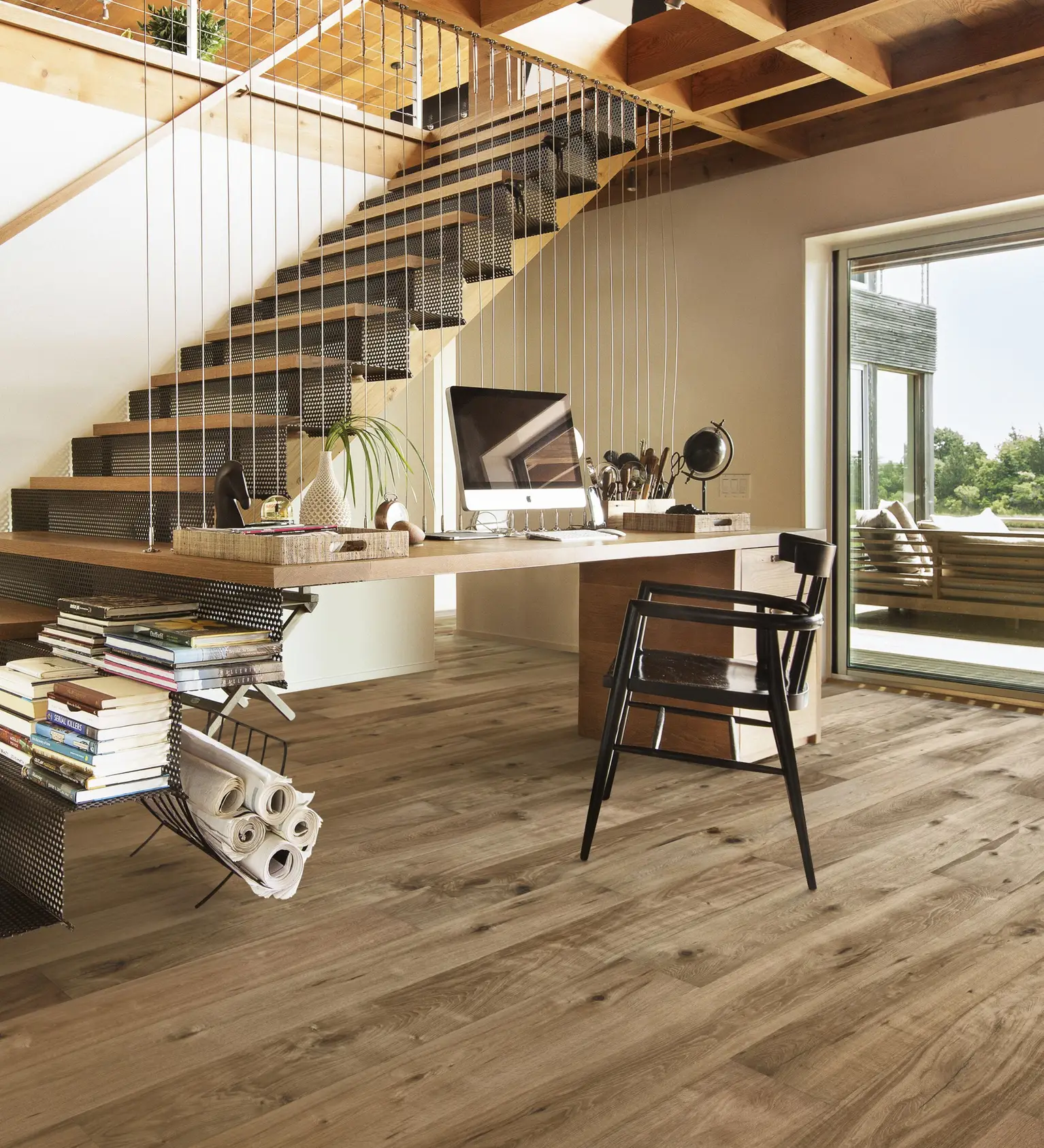 A modern home office with a natural wood desk and matching floor, complemented by an open staircase with metal accents and large windows for natural light.