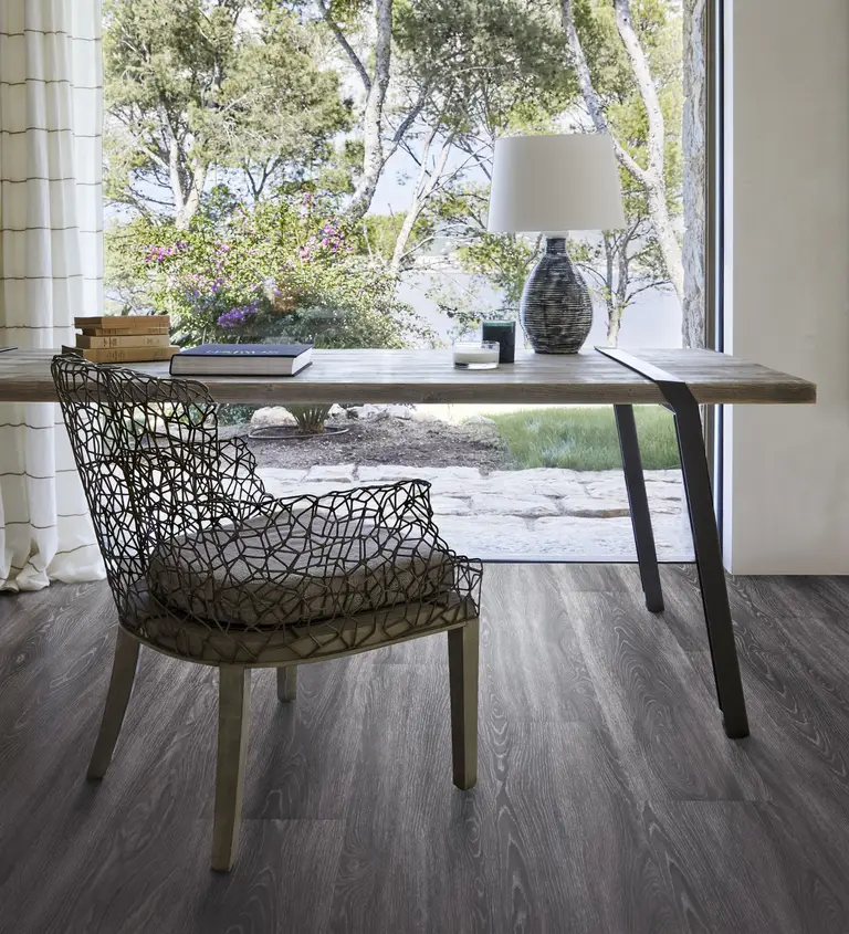 A serene workspace with a rustic wooden desk, gray patterned flooring, and a view of a lush garden through floor-to-ceiling glass doors.