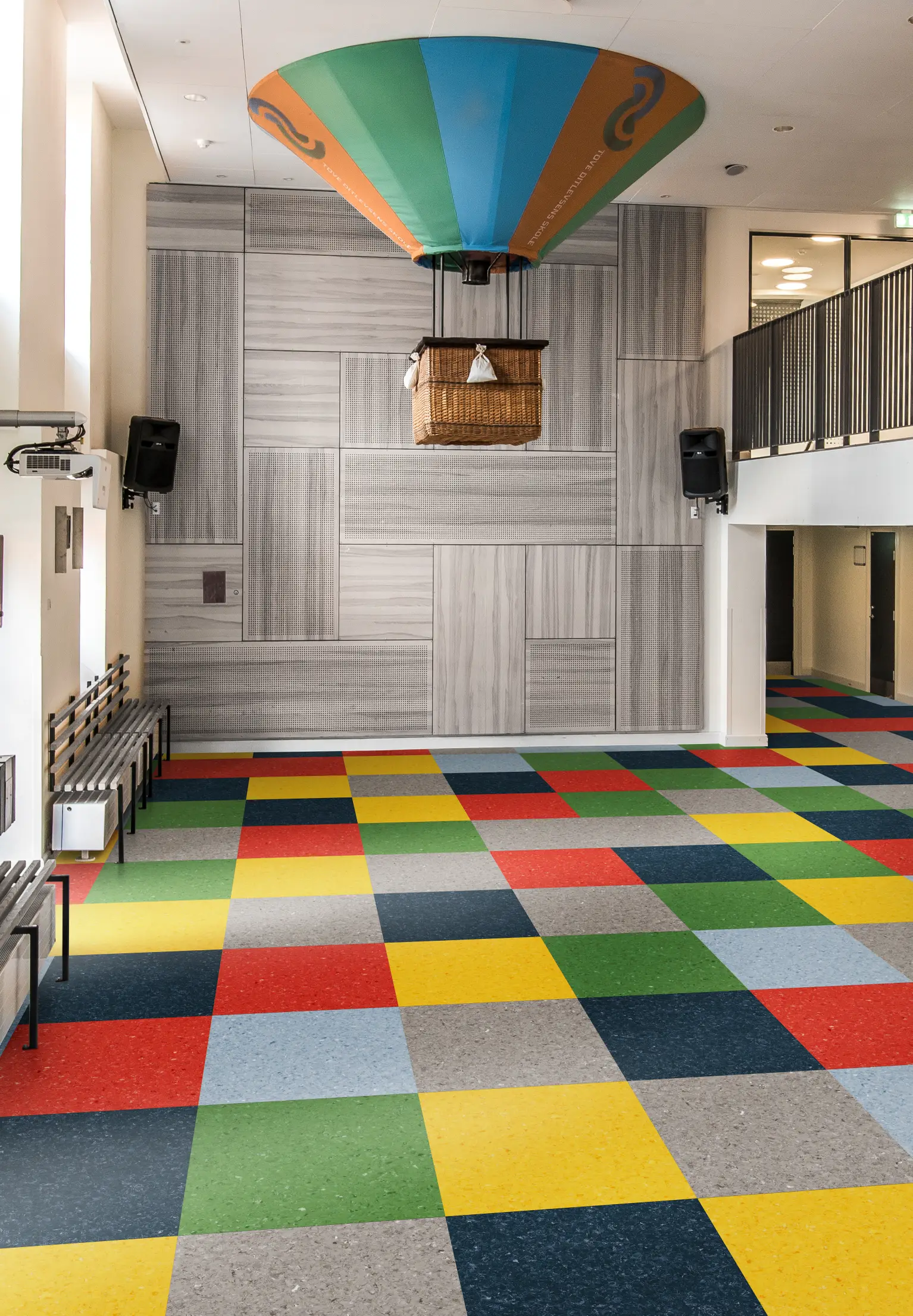 Room with colorful checkered floor tiles, featuring a hot air balloon basket attached to the ceiling, and metallic benches along the walls.
