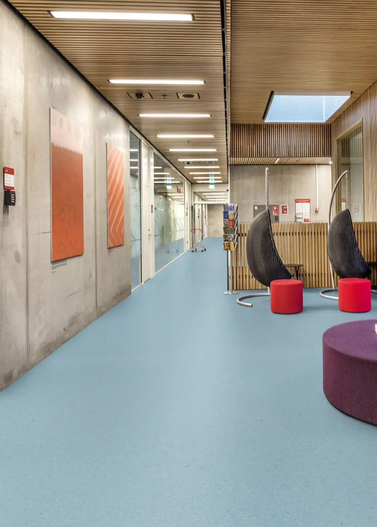 Modern hallway with blue flooring, concrete walls, colorful artwork, and hanging chairs. Bright lighting and a purple ottoman enhance the space.