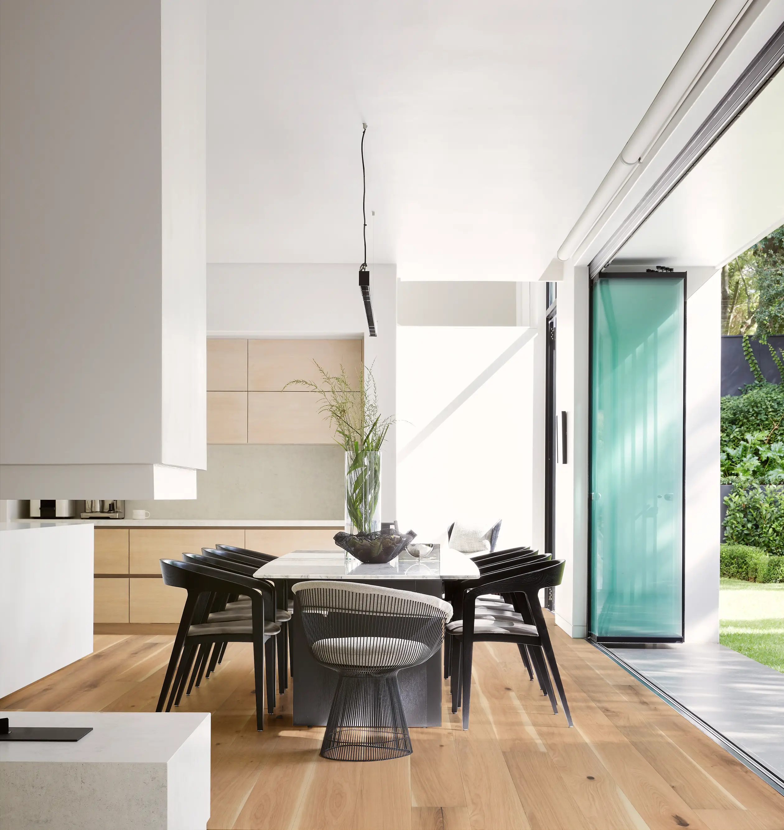Modern dining room with a wooden table, black chairs, and a glass bowl centerpiece. Large glass doors open to a lush garden outside.