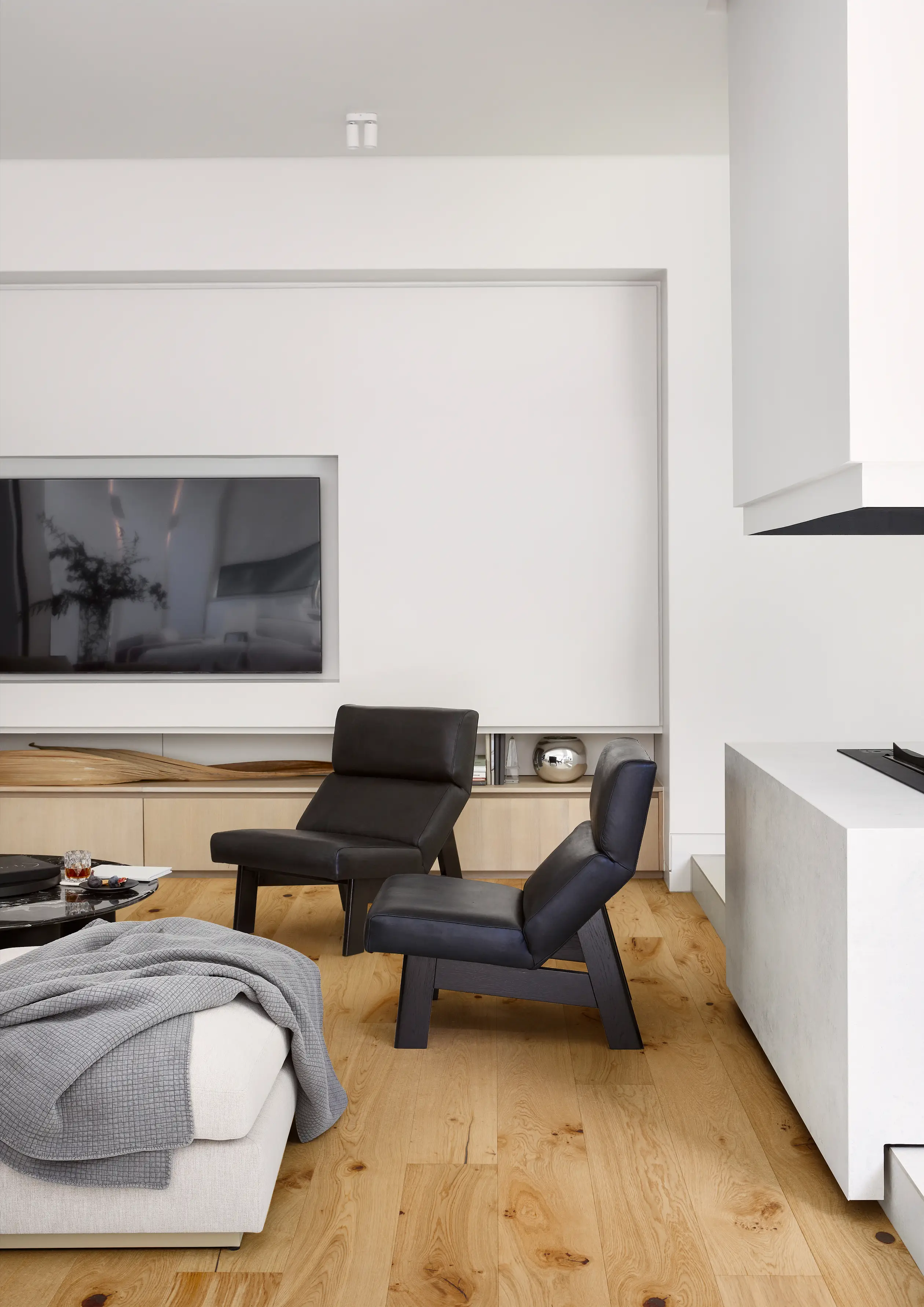Modern living room with two black chairs, a large TV, light wood flooring, and a white sofa with a gray blanket. Minimalist decor.