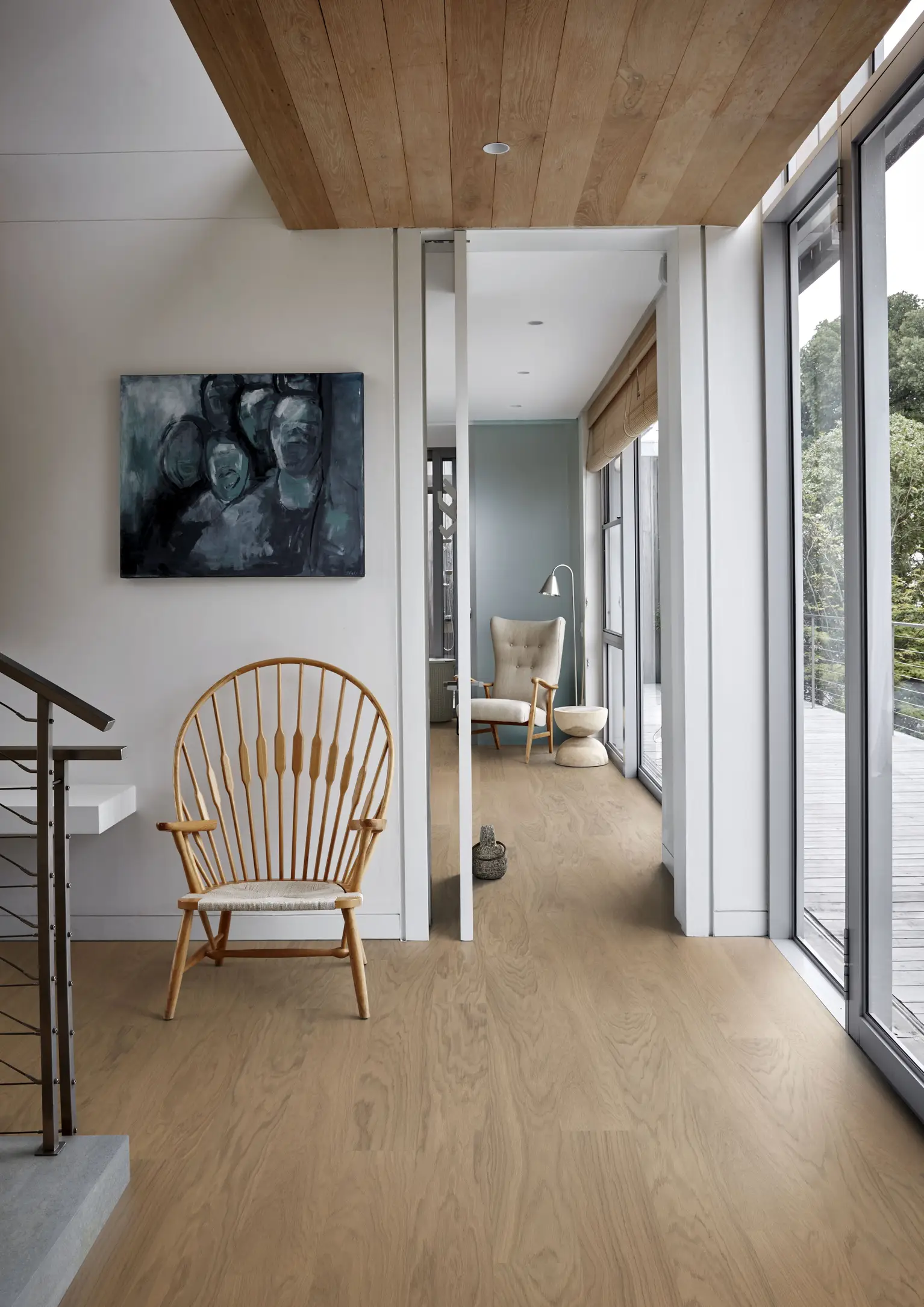 Modern interior with wooden floors, a wicker chair, abstract painting, and large windows overlooking greenery. Cozy nook with a cushioned chair.