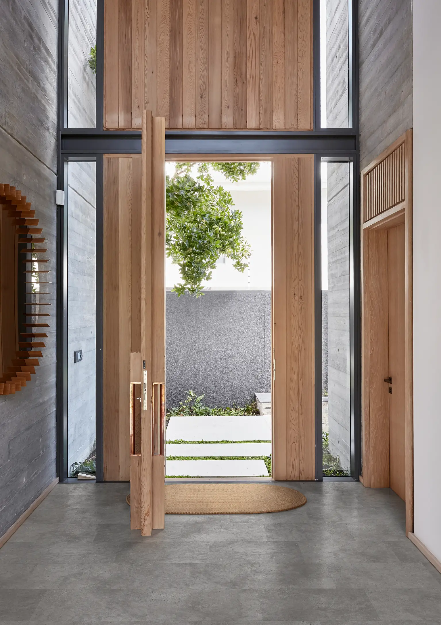 Modern entryway with tall wooden doors, concrete walls, and a view of a small garden outside. A round mirror hangs on the left wall.