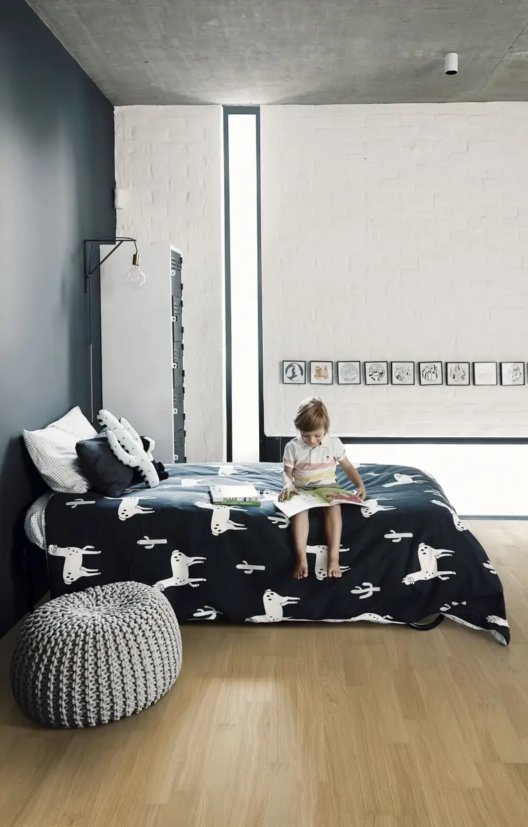 Child sitting on a bed with a dark animal-patterned blanket in a modern bedroom with white walls, wooden floor, and a knitted pouf.