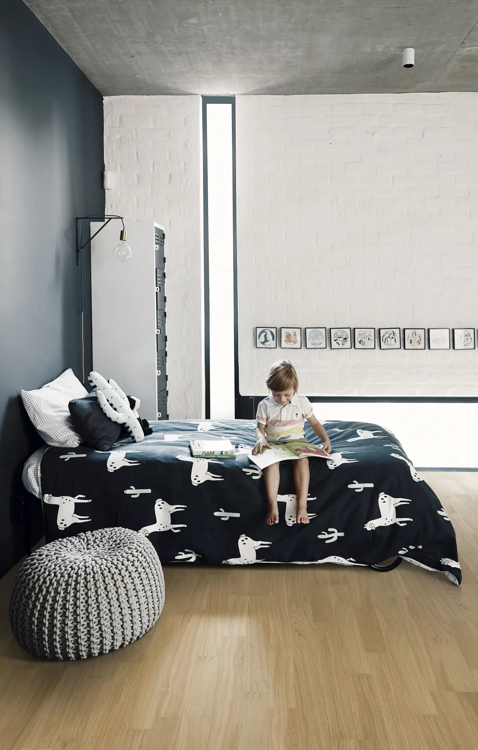 A minimalist children's bedroom with light wooden flooring, dark and white walls, and a child sitting on a bed with playful printed bedding.