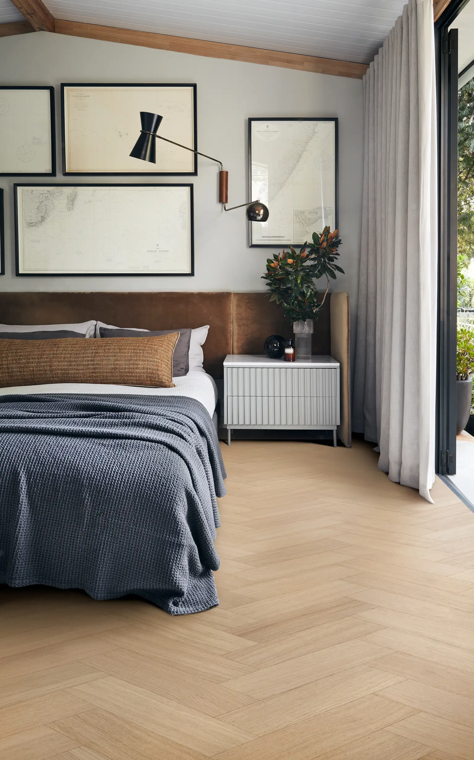 Modern bedroom with a cozy bed, gray blanket, framed maps on the wall, a sleek lamp, and a small plant on a nightstand by a large window.