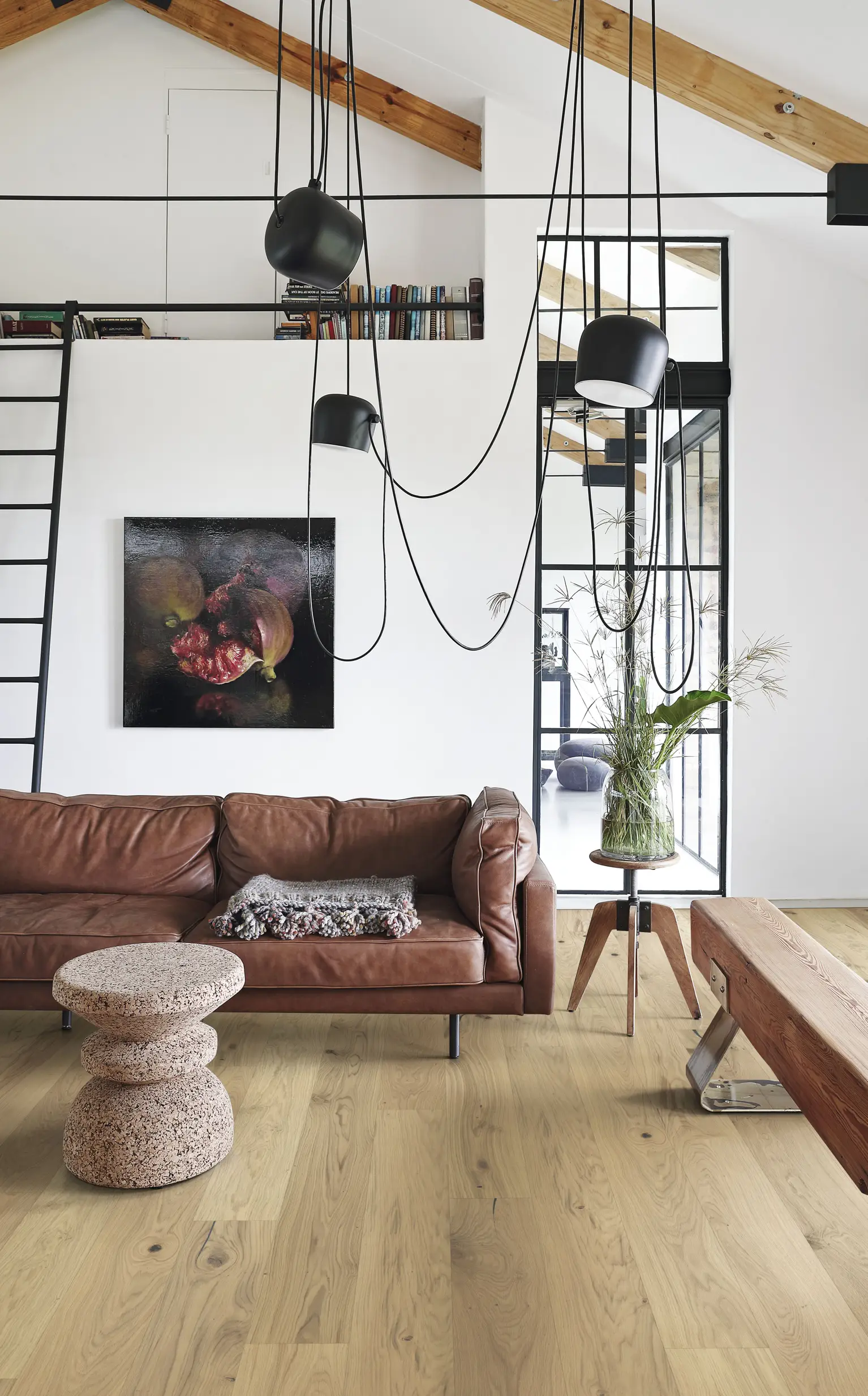Modern living room with a brown leather sofa, abstract art, wooden coffee table, hanging lights, and a ladder leading to a loft bookshelf.