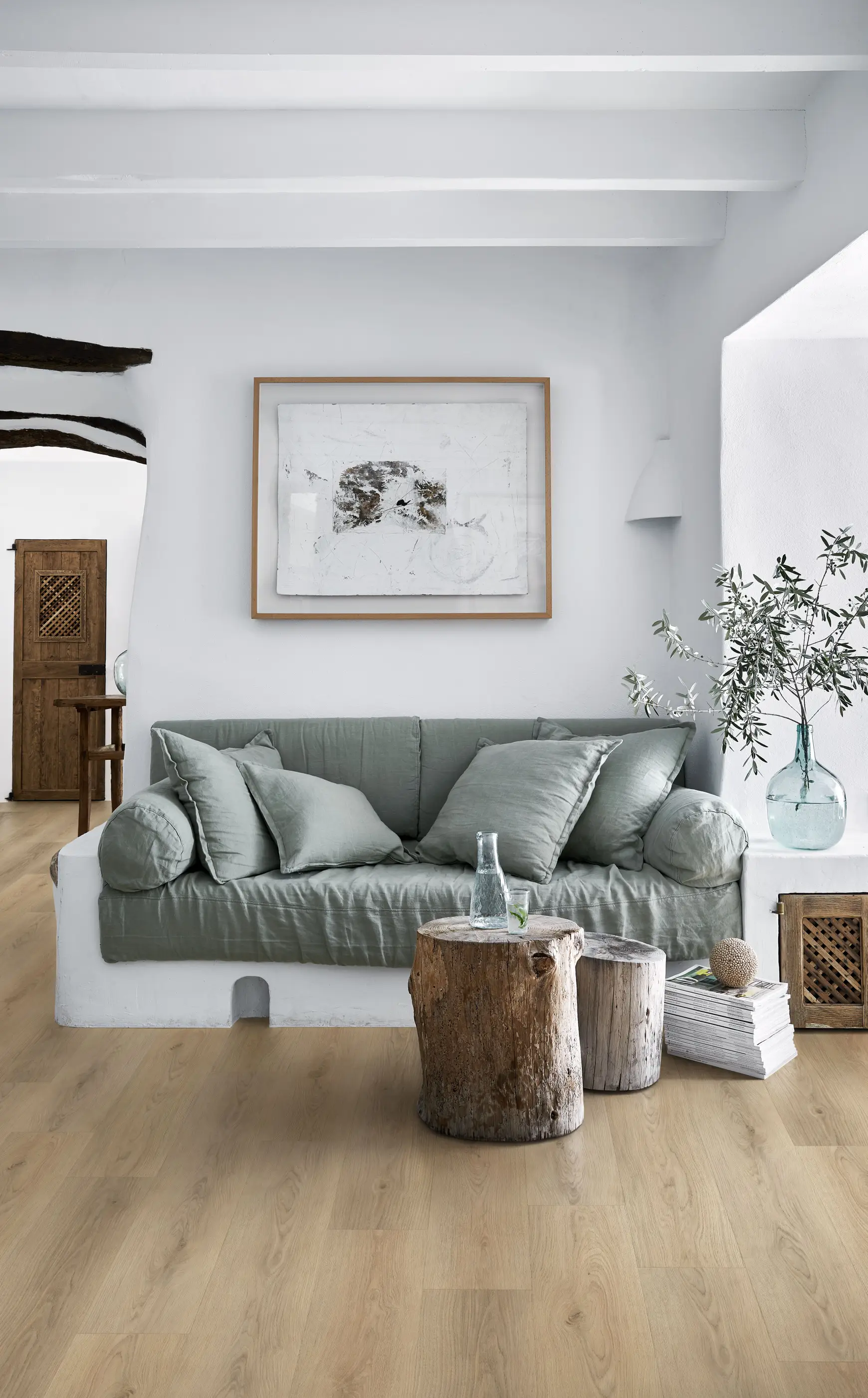 Cozy living room with a green sofa, wooden stump tables, and a framed abstract art piece on a white wall. Natural light and minimalist decor.