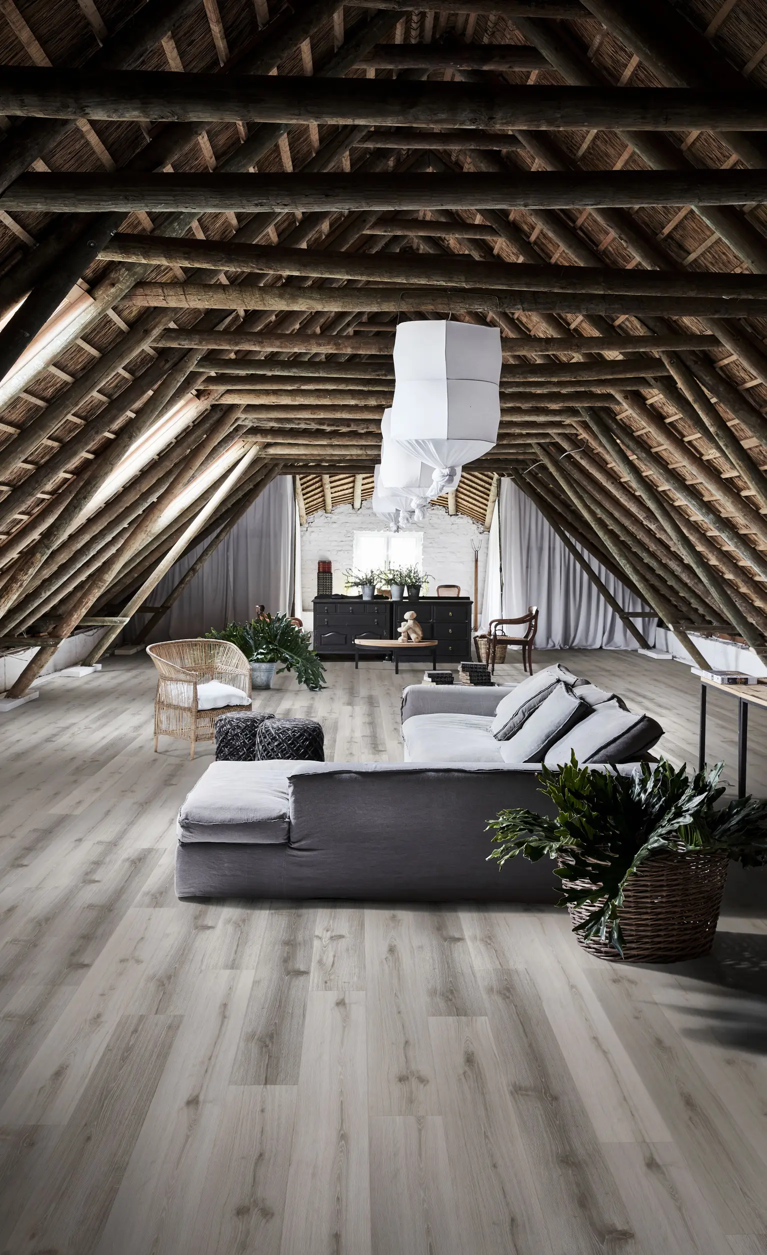Spacious attic with wooden beams, modern gray sofa, wicker chair, plants, and hanging lights, creating a cozy and stylish living area.