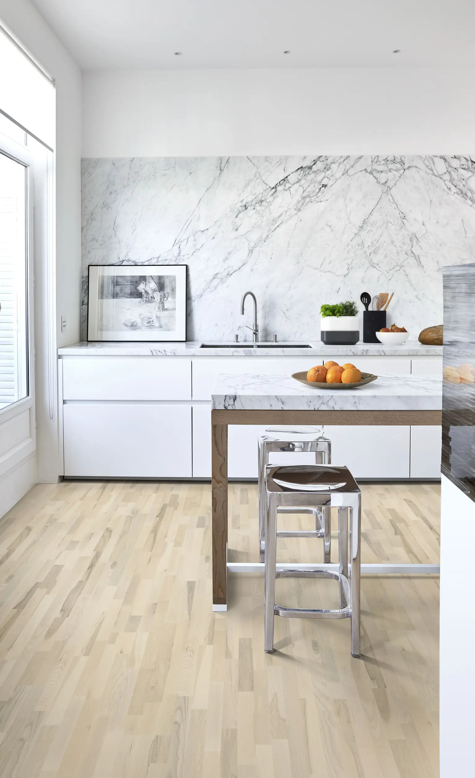 Bright minimalist kitchen with white marble backsplash, marble-topped island, chrome stools, bowl of oranges, and light wood floor.