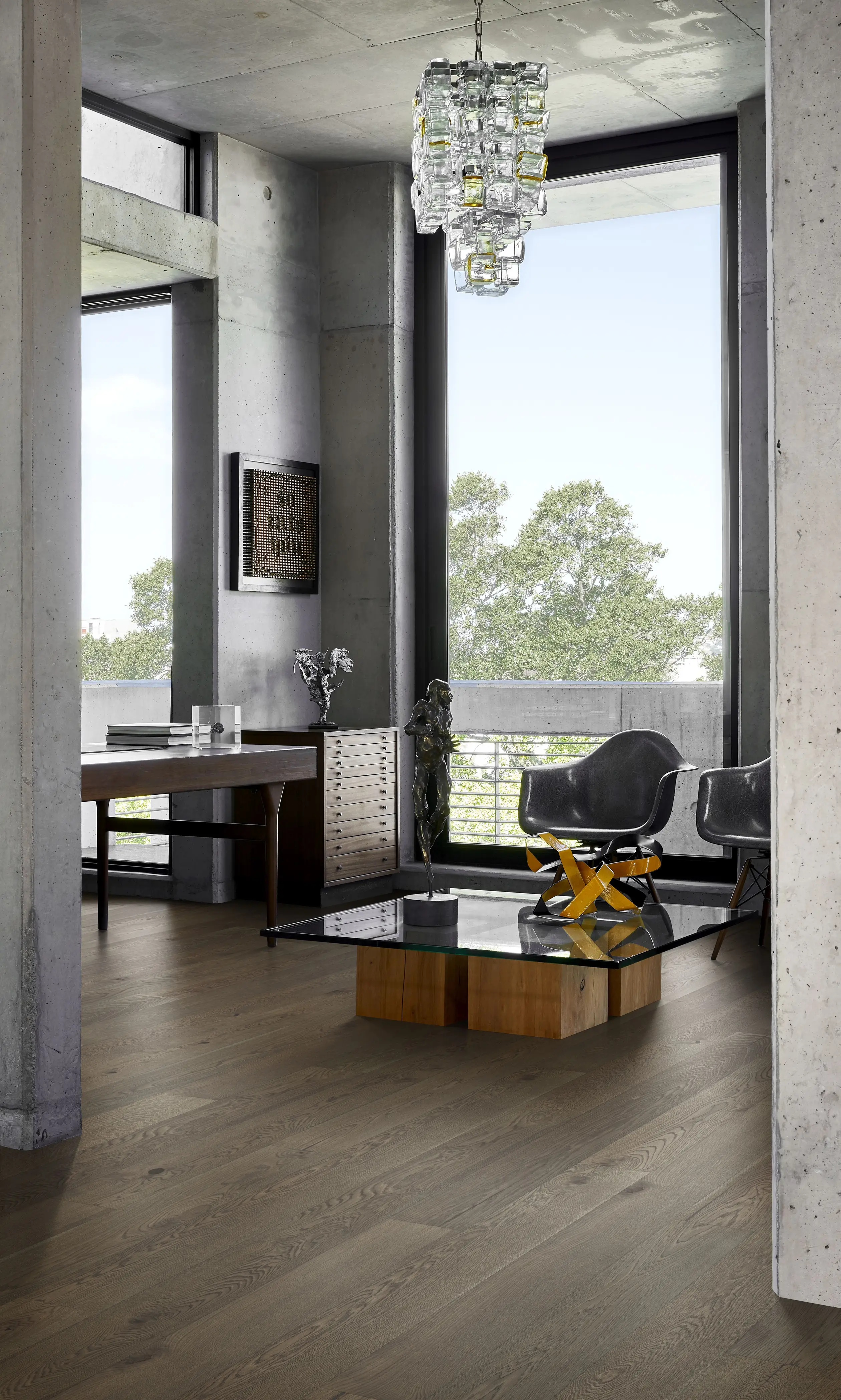 Modern living room with concrete walls, large windows, a glass coffee table, two black chairs, and a cube chandelier. Trees visible outside.