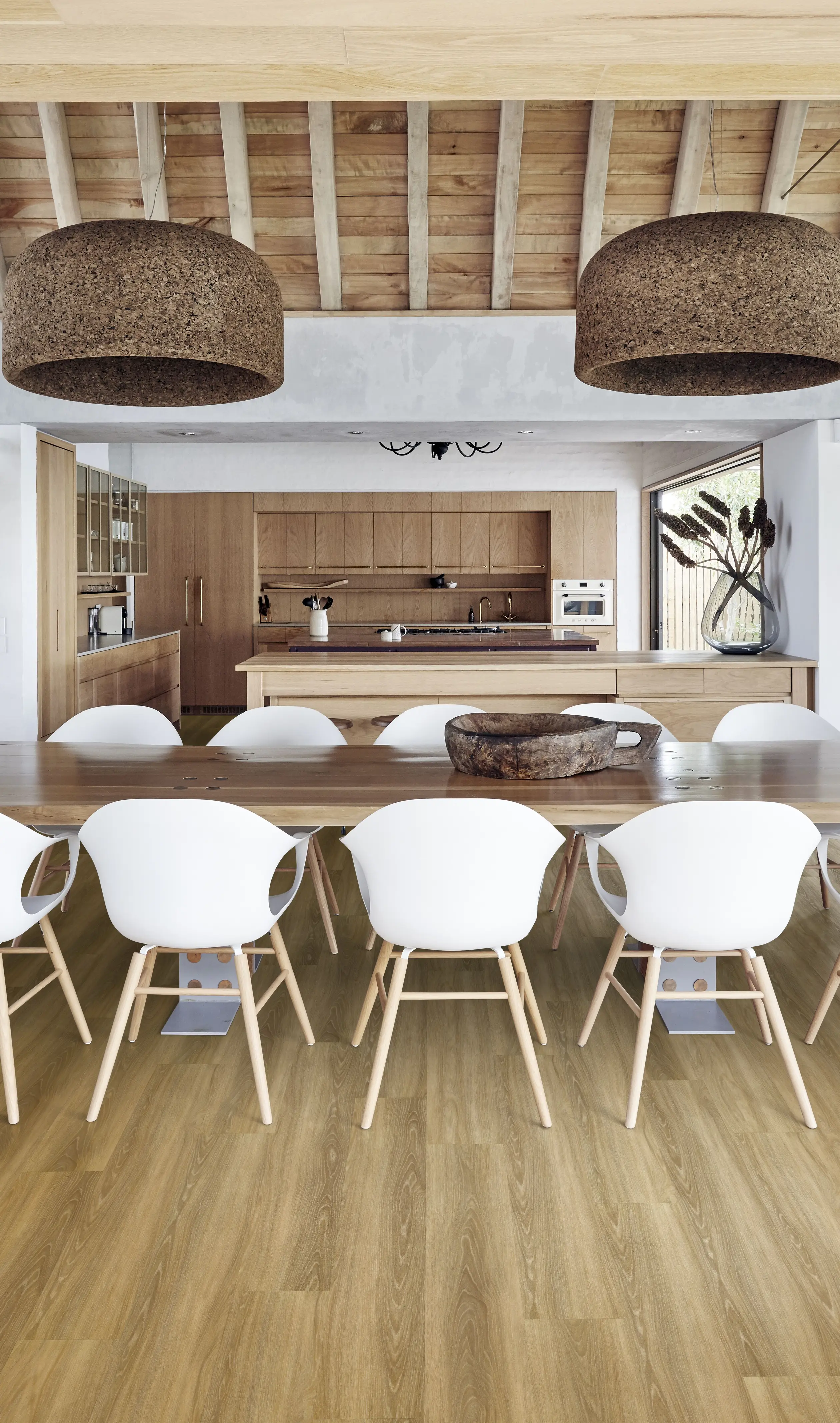 Modern kitchen with wooden elements, featuring a long dining table, white chairs, large pendant lights, and a spacious island with appliances.