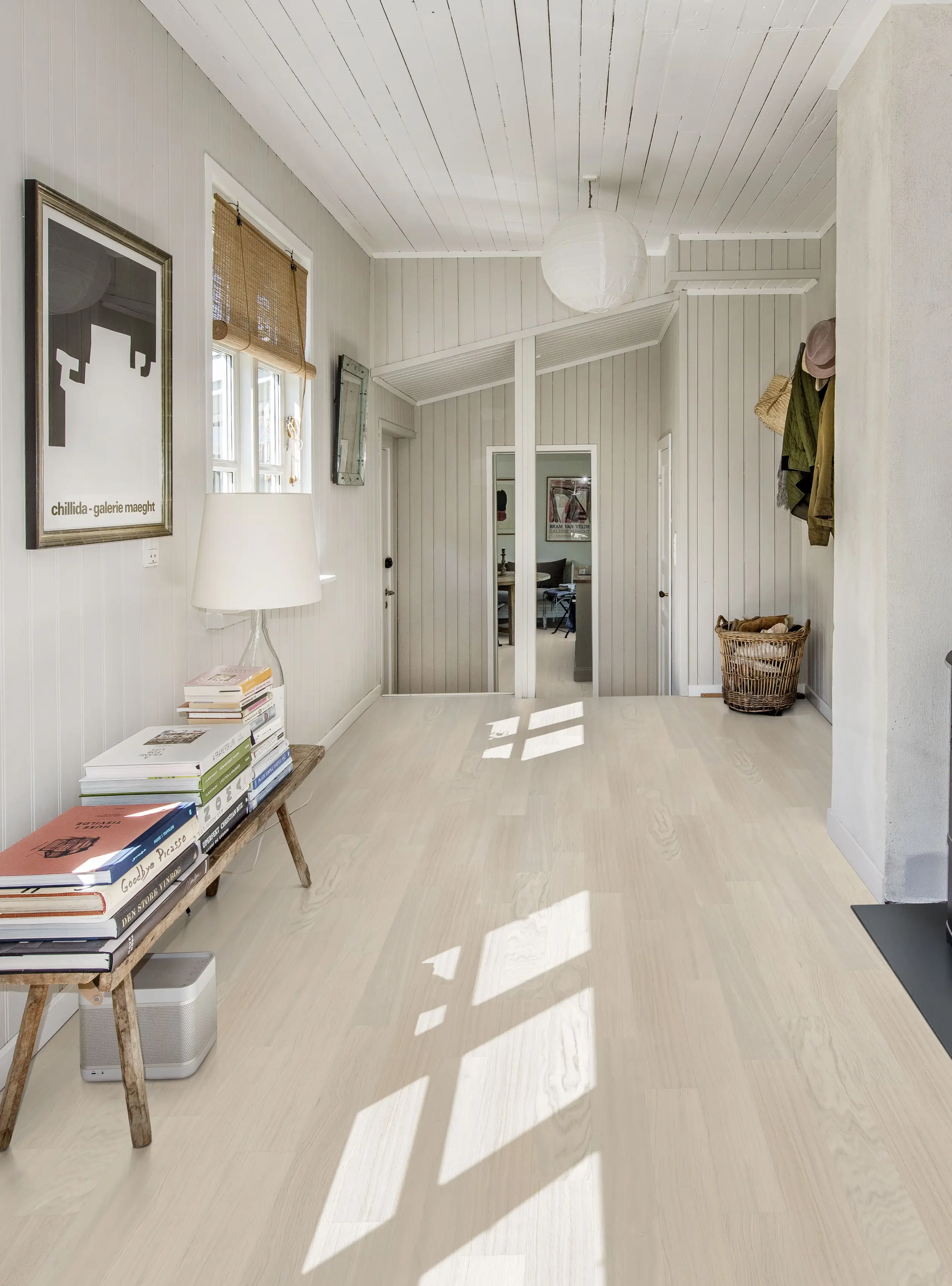 A bright, minimalist hallway with light wood floors, white walls, a bench with stacked books, framed art, and a paper lantern ceiling light.