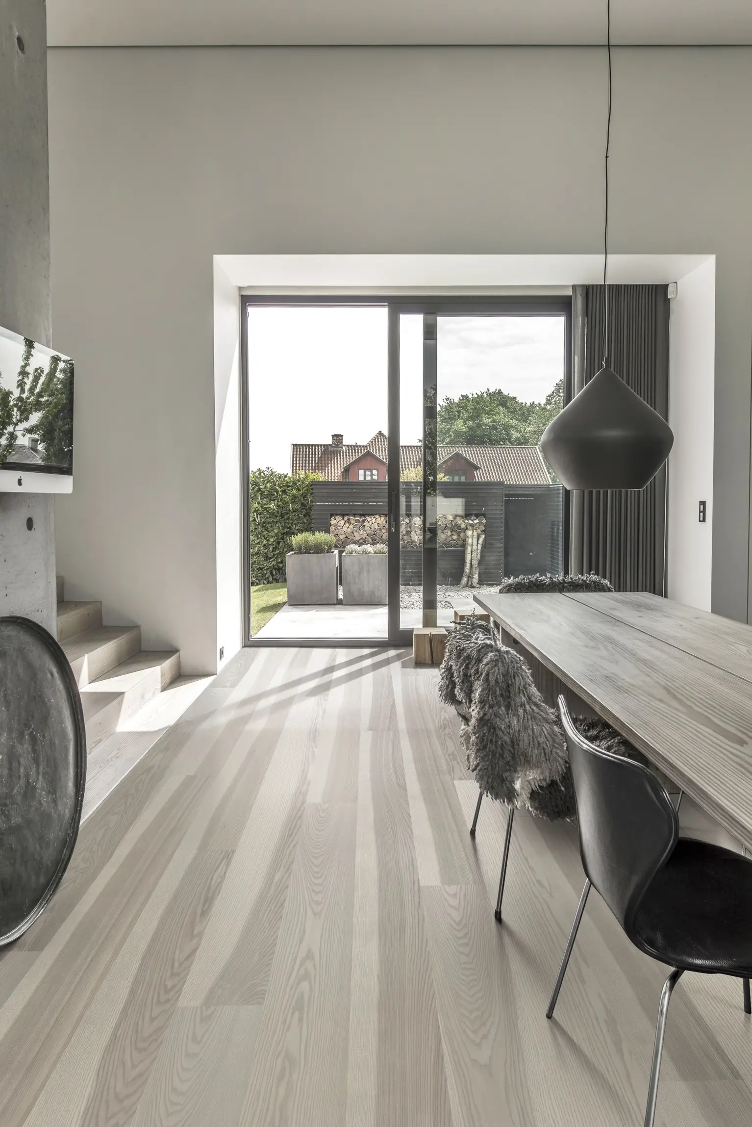 Modern dining area with a wooden table, black chairs, a pendant light, and large glass doors opening to a patio with greenery outside.