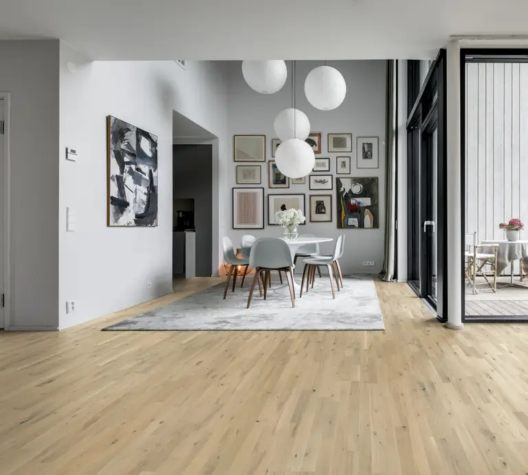 Modern dining area with a light wood floor, round table, gray chairs, and pendant lights. Art-filled wall and large windows offering natural light.