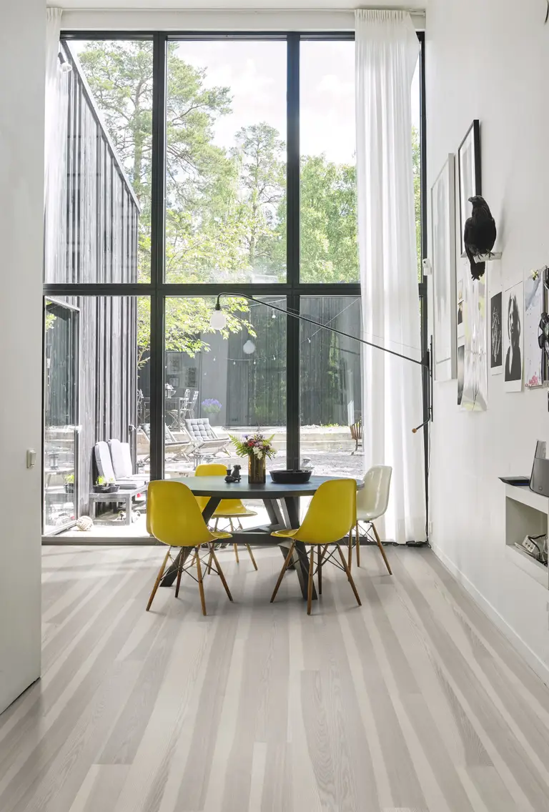 Modern dining area with a round table, yellow chairs, and large windows overlooking a patio and greenery. White curtains and light wood floors.