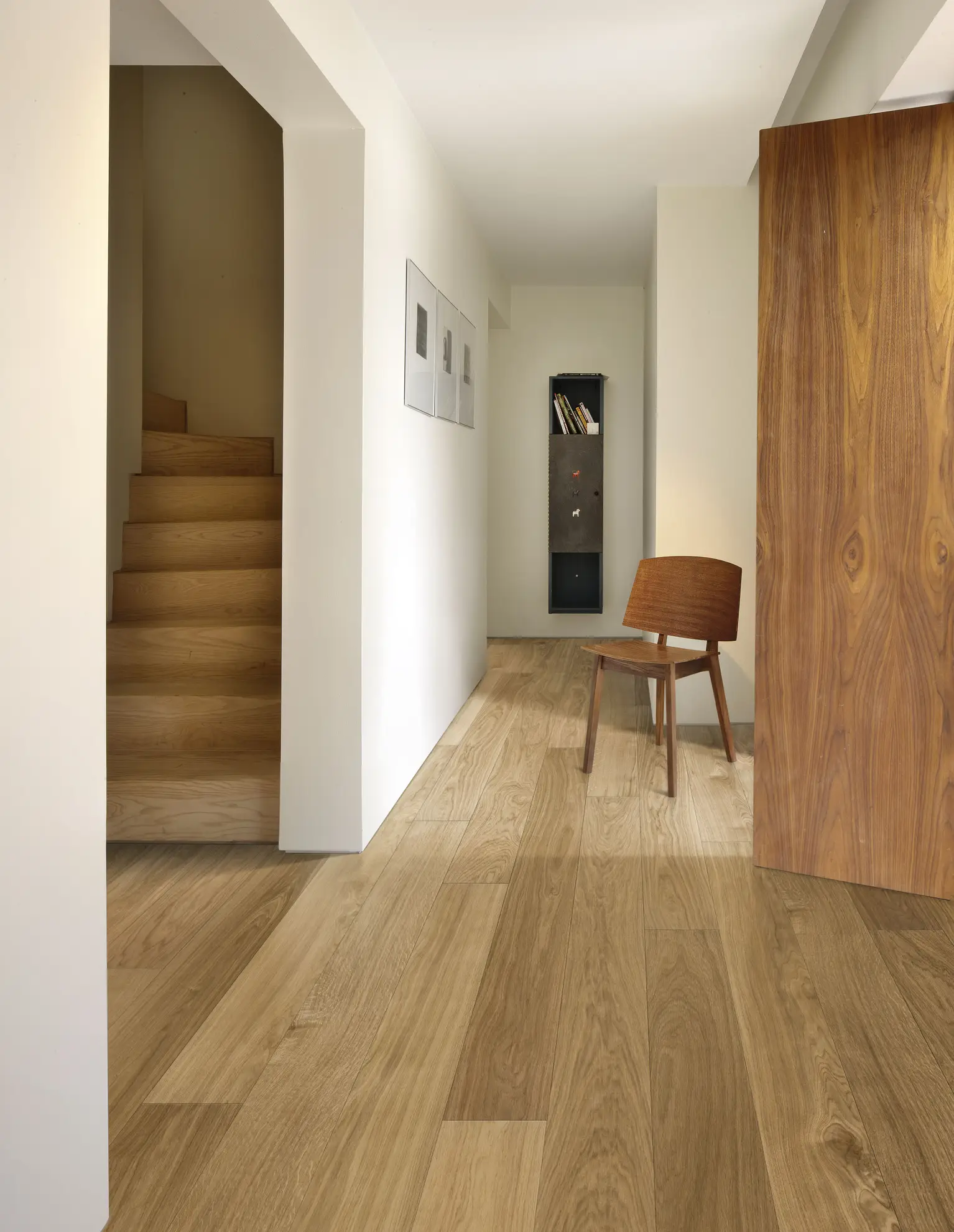 Minimalist hallway with light wood flooring, a wooden chair, staircase on the left, and a small shelf in the background. Natural light illuminates the space.