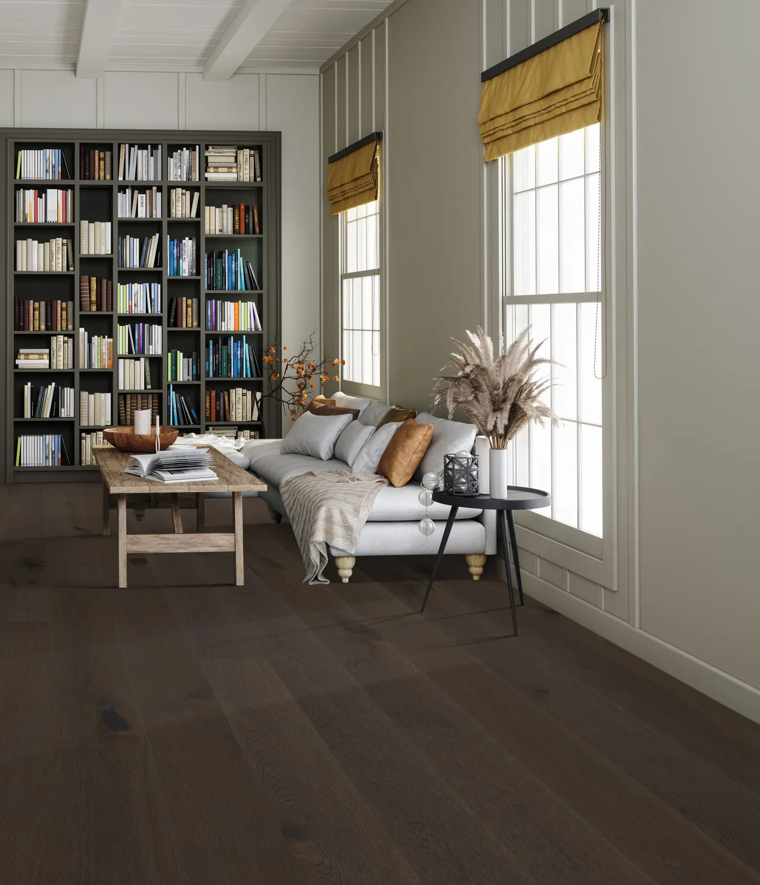 A cozy living room with a wooden floor, a gray sofa, decorative pillows, and a rustic coffee table. A bookshelf fills the wall near tall windows.