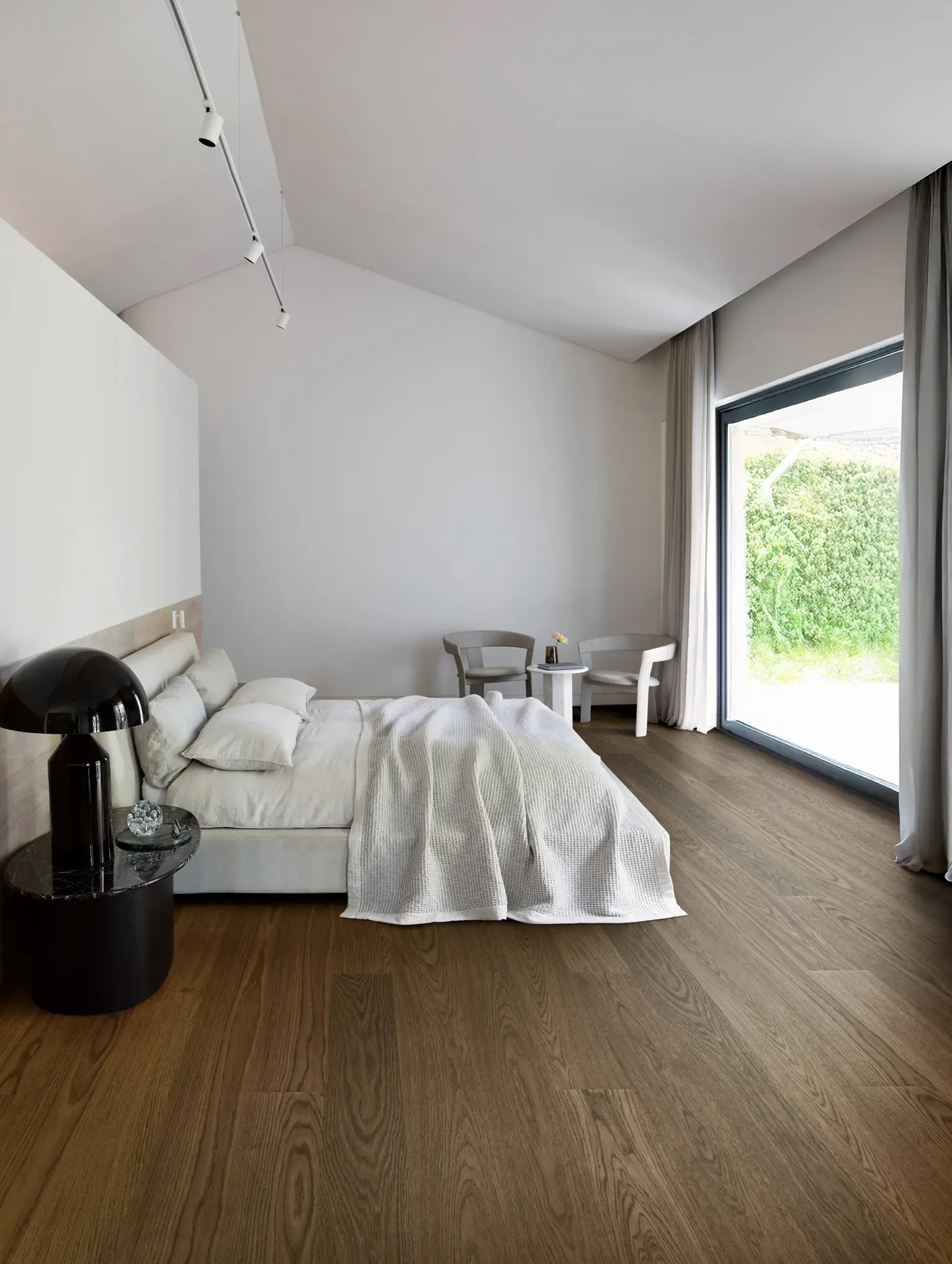 Minimalist bedroom with a large bed, white bedding, two chairs, a small table, and a floor-to-ceiling window overlooking greenery.