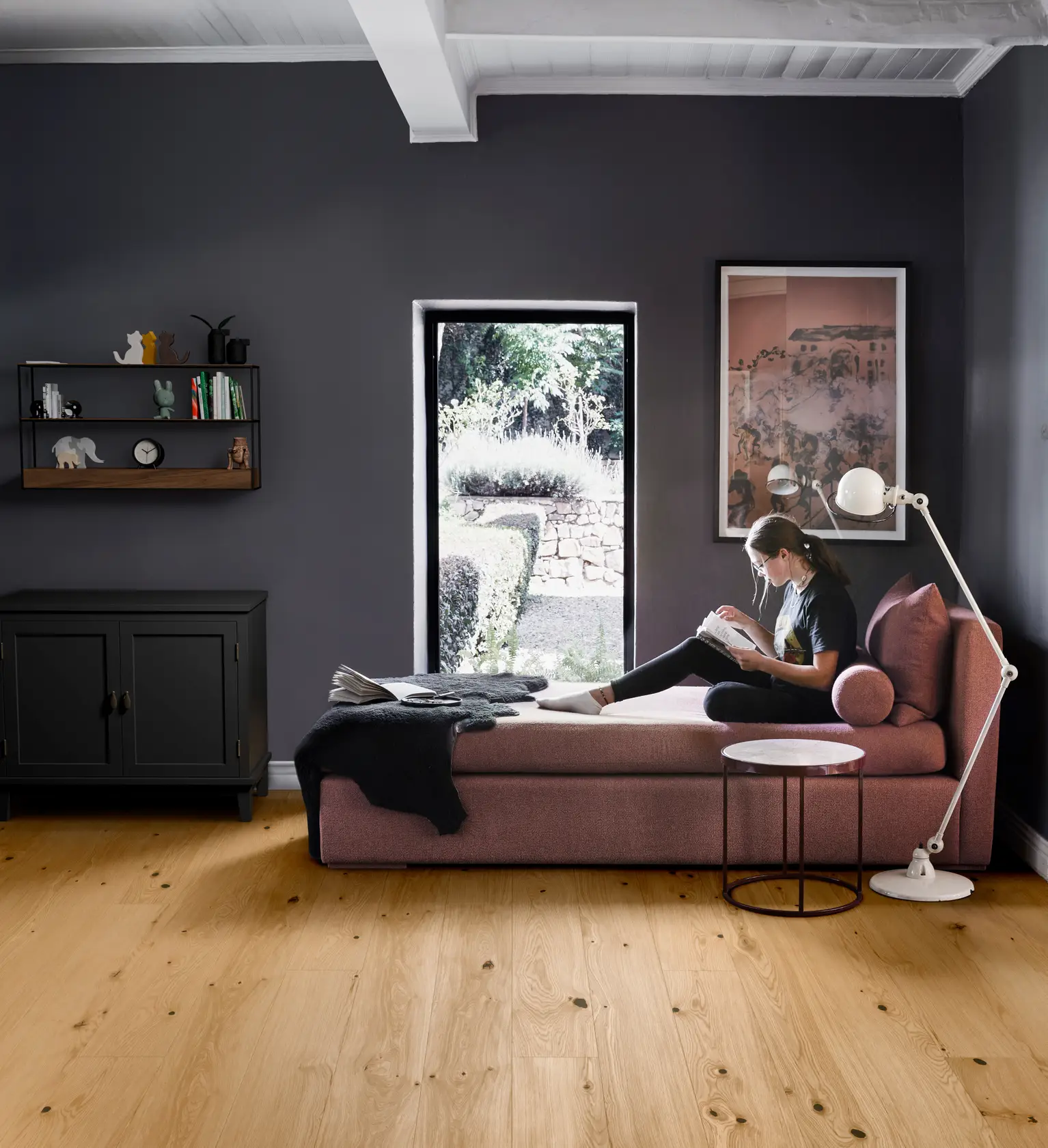 Woman reading on a pink chaise lounge in a modern room with dark walls, wooden floor, and a window view of a garden.