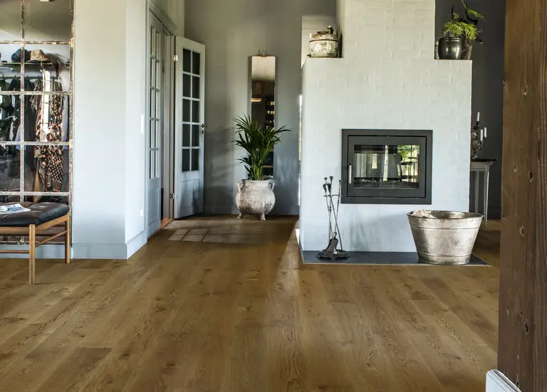A cozy living room with wooden flooring, a white brick fireplace, potted plants, and a large mirror reflecting light from a glass door.
