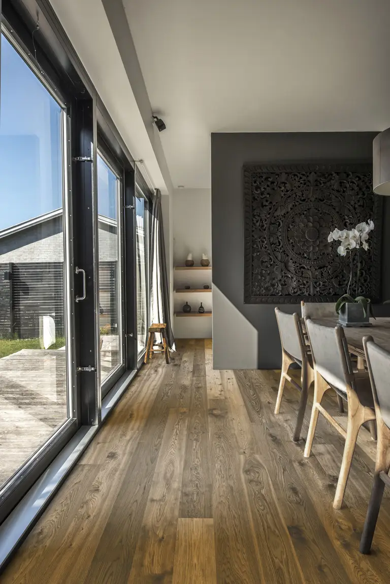 Modern dining area with large windows, wooden floor, and a decorative wall panel. A table with chairs and a potted orchid are visible.