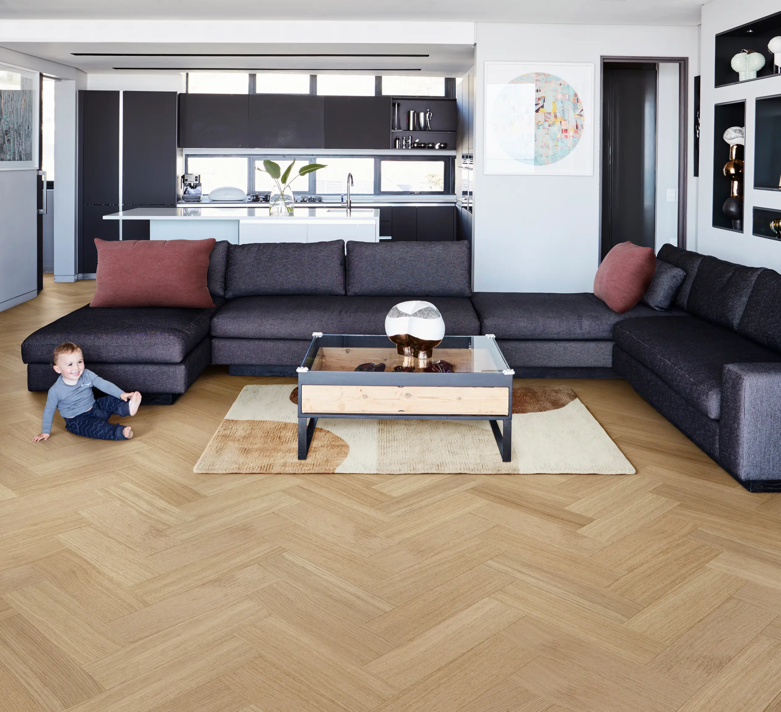Modern living room with a dark gray sectional sofa, glass coffee table, and light wood flooring. A child sits on the floor near the sofa.