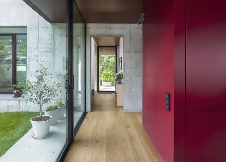 Oak Dublin. A sleek hallway with light oak flooring, a bold red accent wall, and large glass windows overlooking greenery.