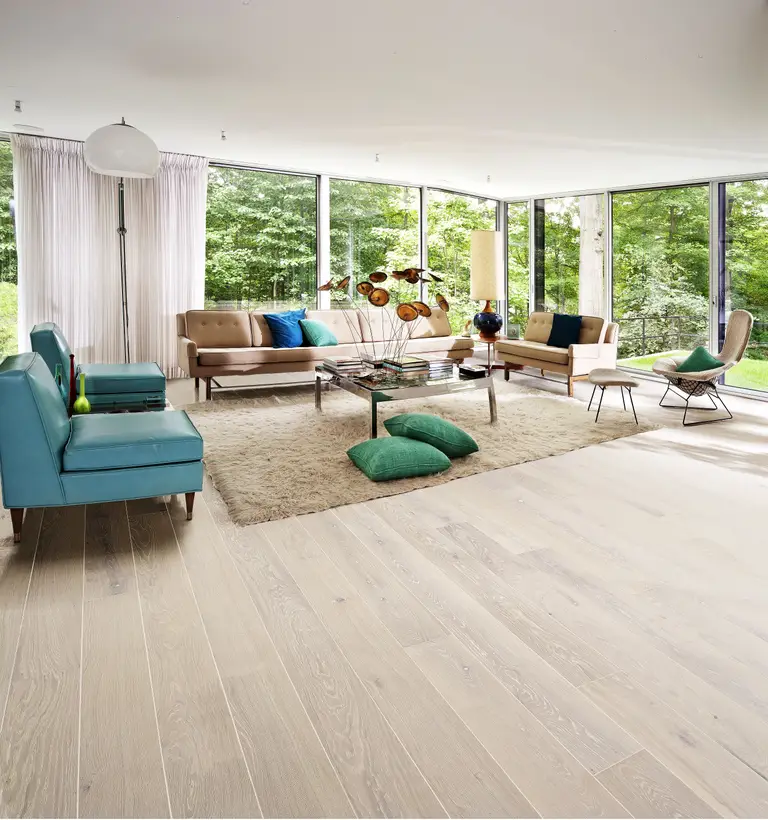 Modern living room with large windows, light wood flooring, blue and beige sofas, a shag rug, and green cushions. Forest view outside.