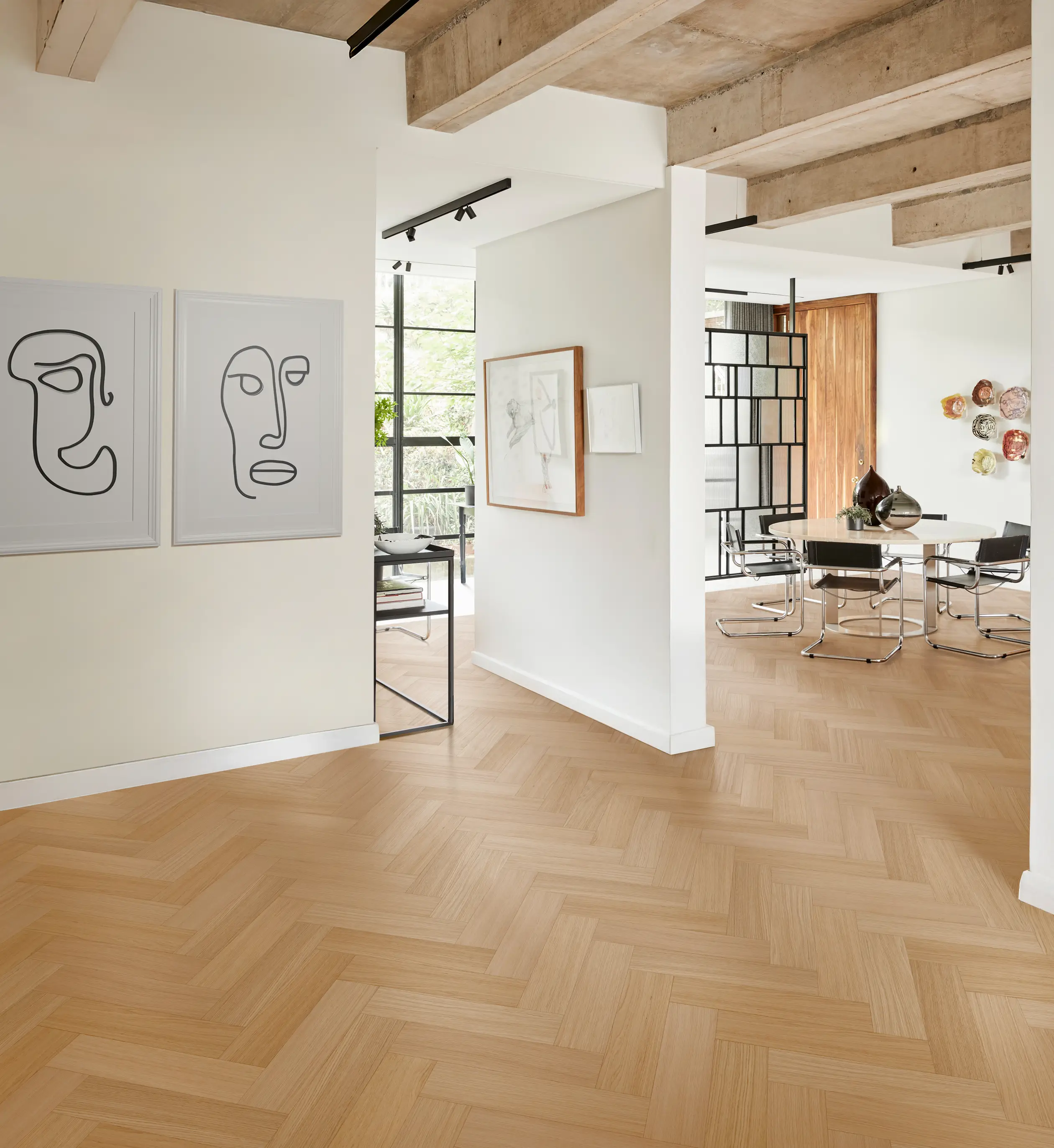 Modern interior with light wood herringbone floor, abstract face art on walls, and dining area featuring black chairs and a wooden table.