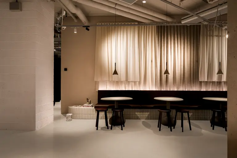 Modern cafe interior with round tables, dark stools, and pendant lights. Soft, warm lighting and draped curtains create a cozy atmosphere.