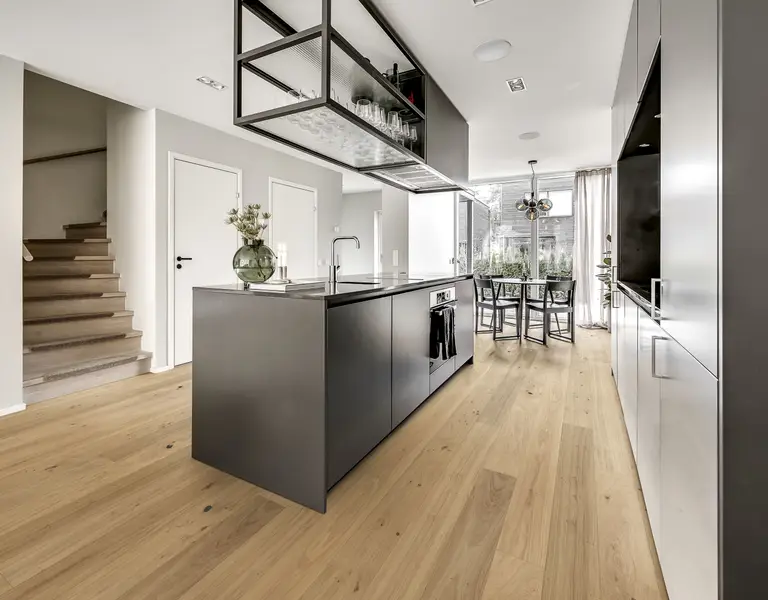 Modern kitchen with a large black island, wooden floors, and sleek cabinets. A dining table is near the window, and stairs lead to another level.