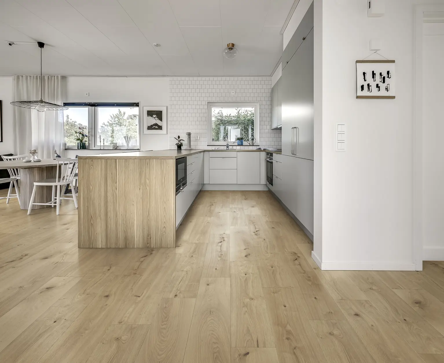 Modern kitchen with light wood flooring, a central island, white cabinets, and large windows. Minimalist decor and natural light enhance the space.