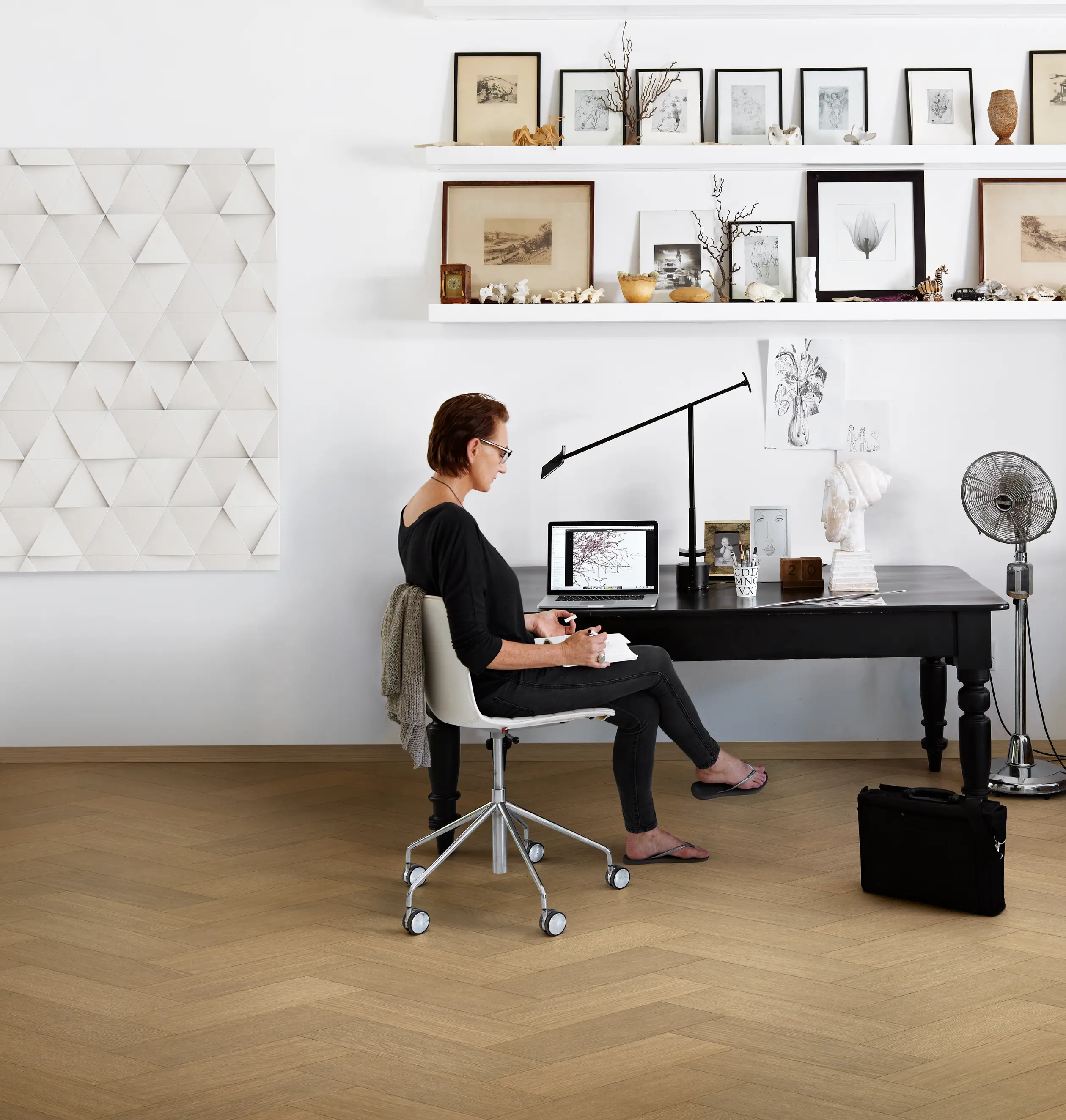 Person seated at a desk in a stylish room with art on shelves, working on a tablet. A geometric wall art piece and fan are visible.