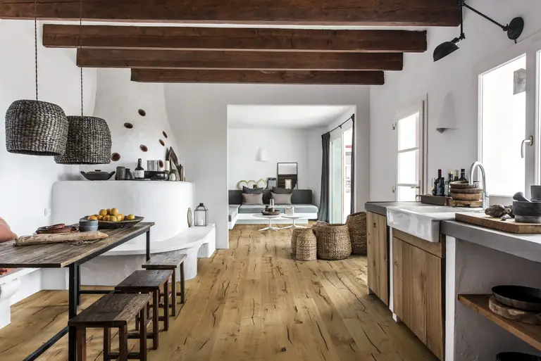 Rustic kitchen with wooden beams, a long dining table, woven pendant lights, and a cozy living area with natural light and neutral tones.