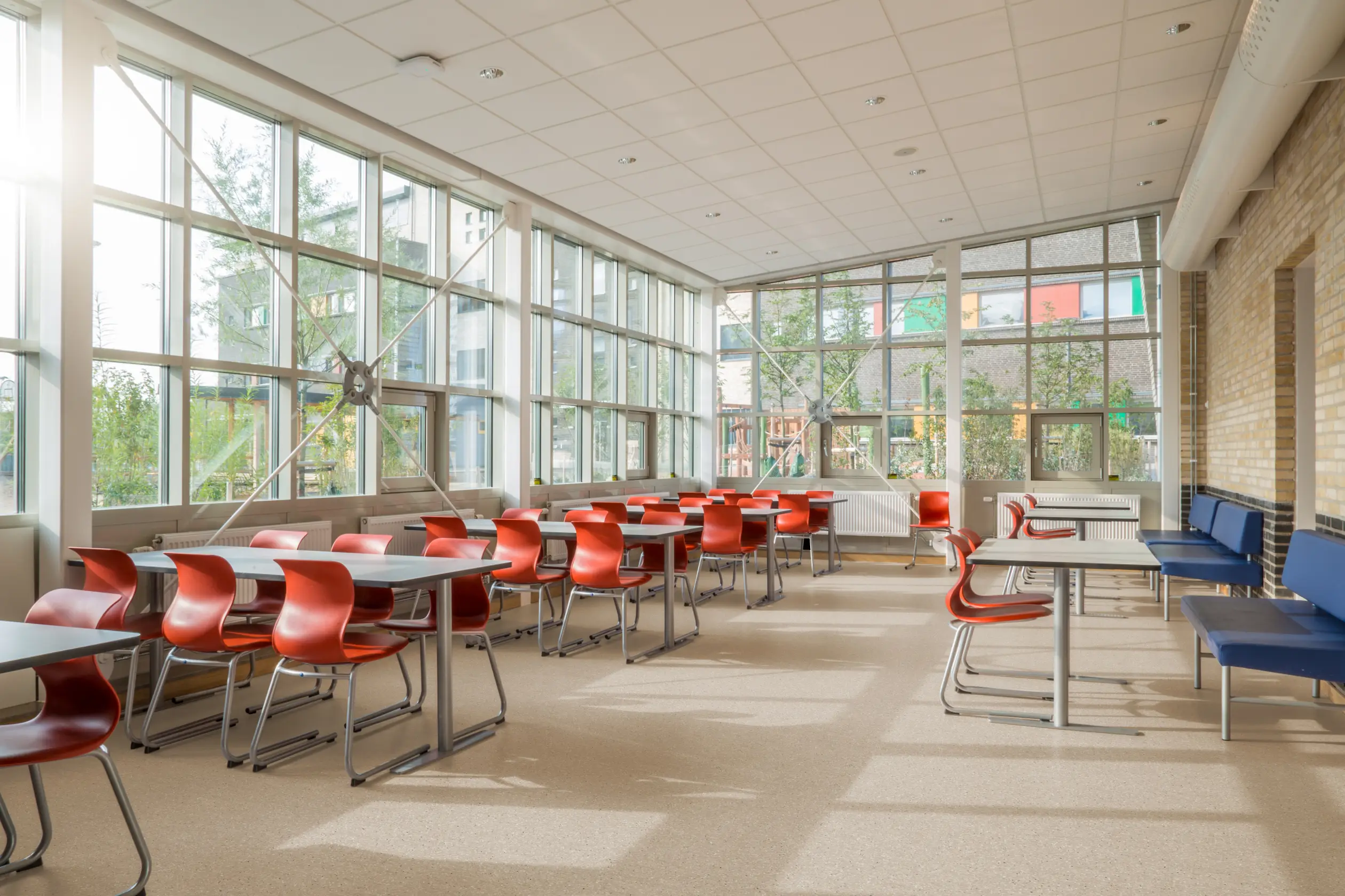 A bright, empty classroom with red chairs, wooden desks, and large windows allowing natural light to flood the space.