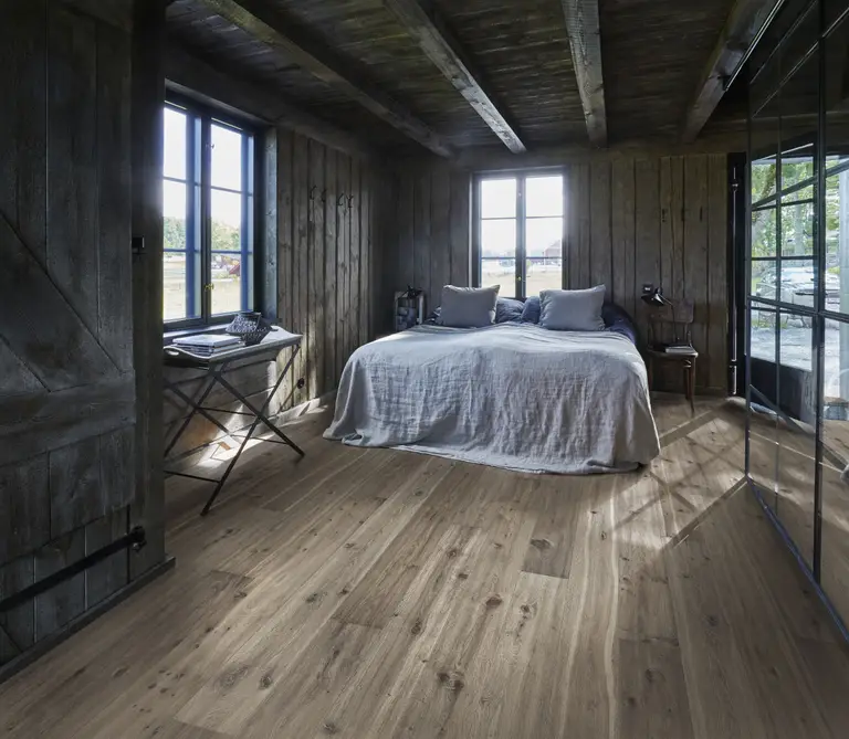Cozy rustic bedroom with wooden walls and floor, featuring a double bed with gray bedding, large windows, and soft natural light.