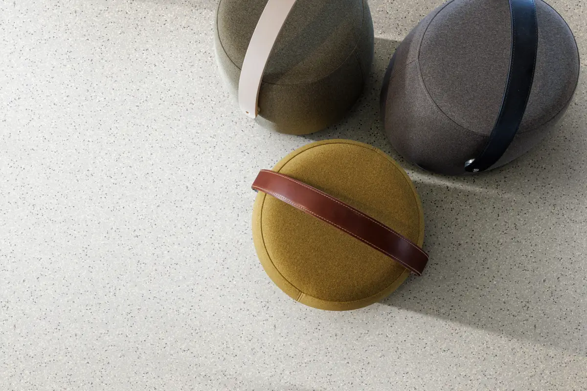 Three modern, round fabric stools with leather handles in brown, beige, and dark gray, placed on a light speckled surface.