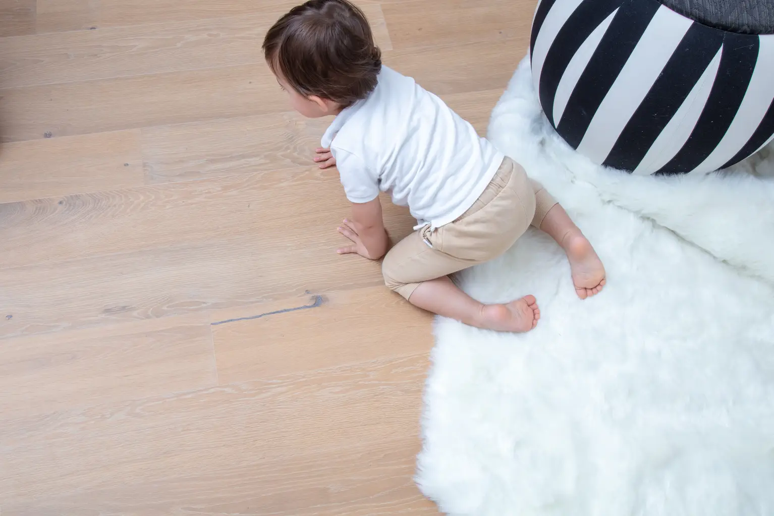 A baby in a white shirt and beige pants crawls on a wooden floor, partially covered by a fluffy white rug and a striped cushion.