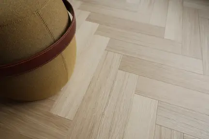 Close-up of mustard yellow round pouf with brown leather strap on light wood herringbone parquet floor.