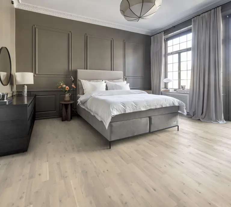 Modern bedroom with a large bed, gray walls, wooden floor, and a big window with gray curtains. A dresser and round mirror are on the left.