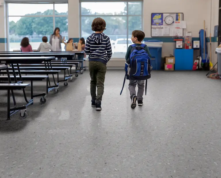 Two children with backpacks walk through a school cafeteria. Tables and a large window are visible, with more children and an adult in the background.