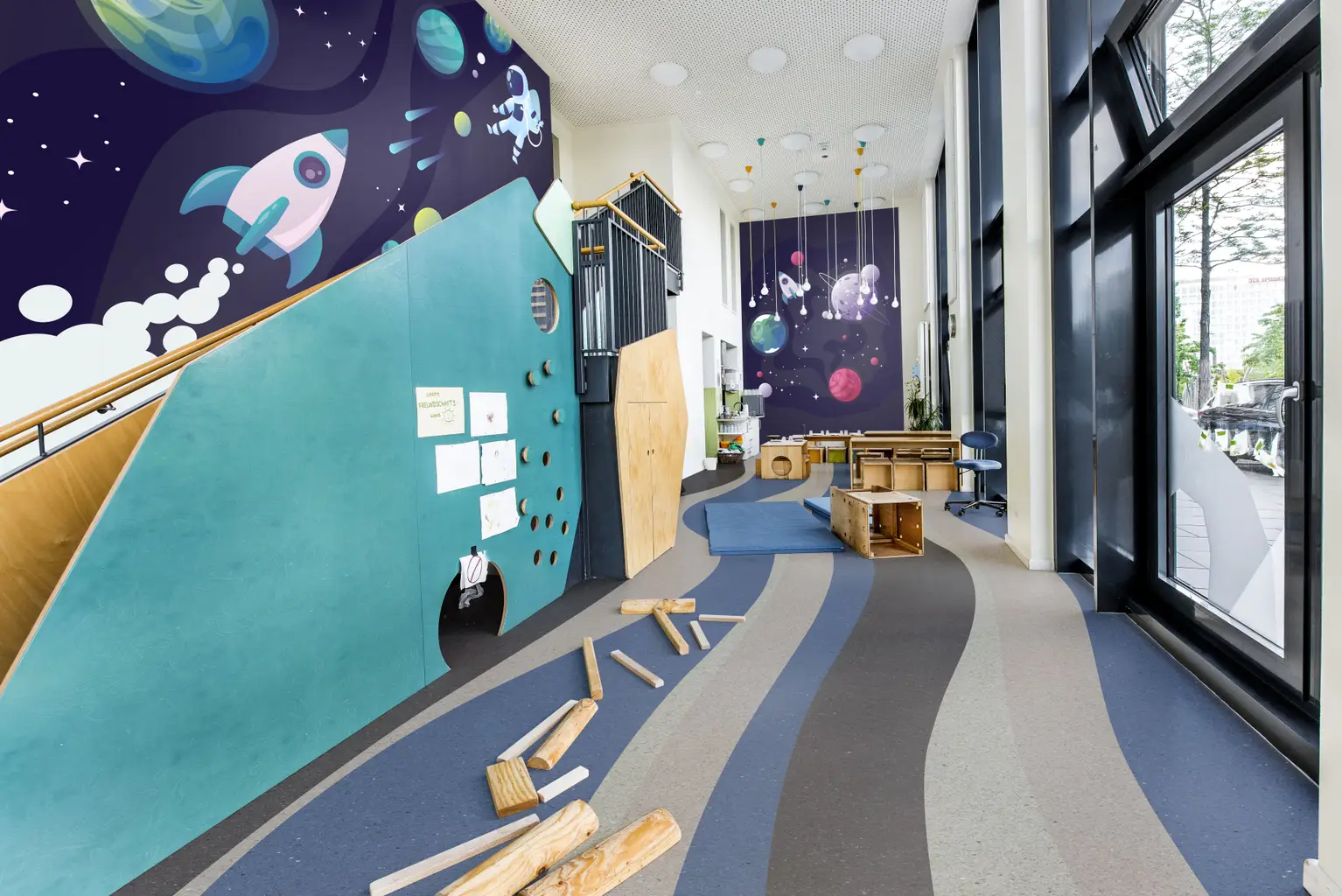 Children's play area with a space-themed mural, climbing wall, and scattered wooden blocks on a striped floor. Large windows on the right.