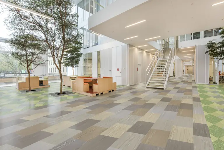 Spacious modern atrium with checkered flooring, indoor trees, wooden seating, and a white staircase leading to an upper level.