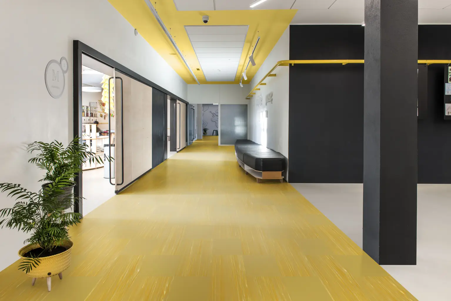 Modern office hallway with yellow flooring, black and white walls, ceiling lights, glass doors, a bench, and a potted plant.