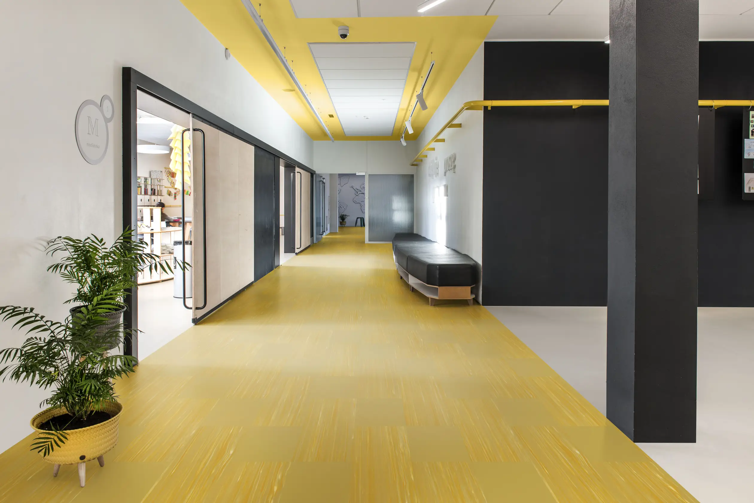 Modern office hallway with yellow flooring, black and white walls, ceiling lights, glass doors, a bench, and a potted plant.