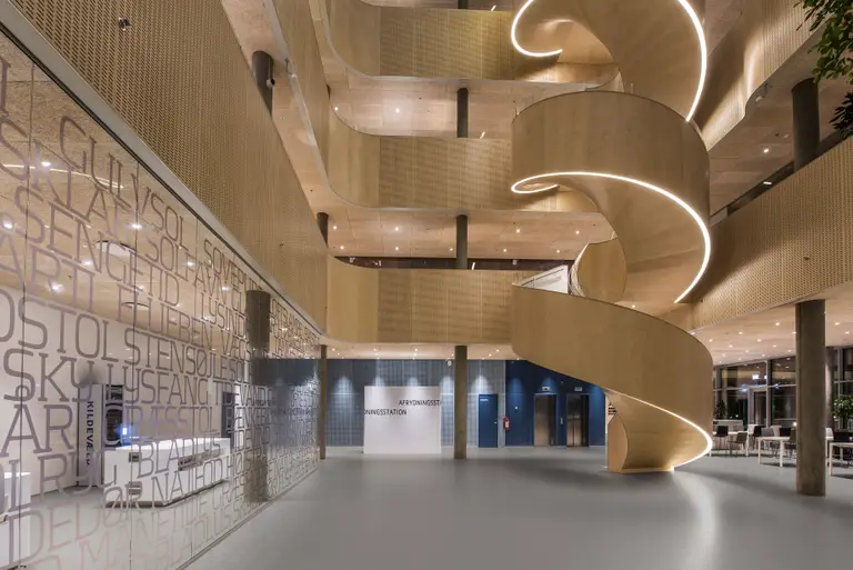 Modern atrium with a large, elegant spiral staircase, illuminated by soft lighting. Walls feature glass and artistic text designs.