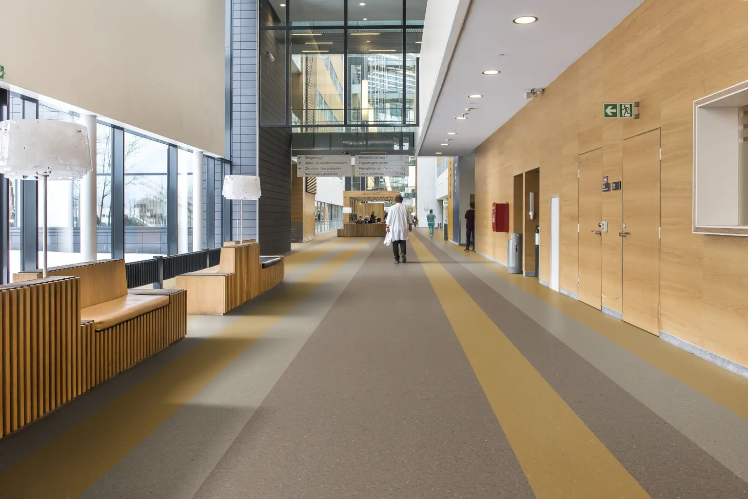 Modern hospital corridor with wood paneling, large windows, and a person in a white coat walking down the hallway.